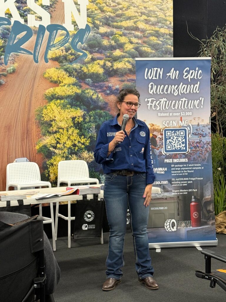 A woman in a blue shirt and jeans speaks into a microphone in front of a promotional banner and table setup at an event.