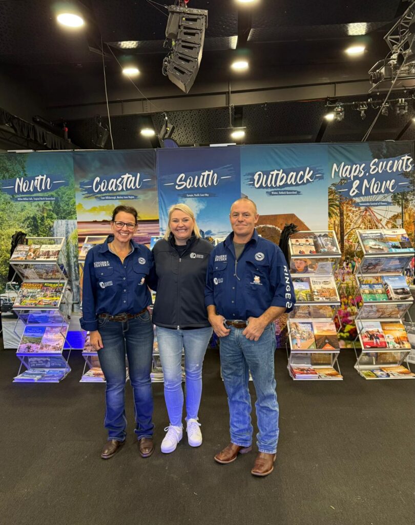 Three people stand smiling in front of travel brochure displays labeled North, Coastal, South, Outback, and Maps, Events & More, inside a well-lit indoor venue.