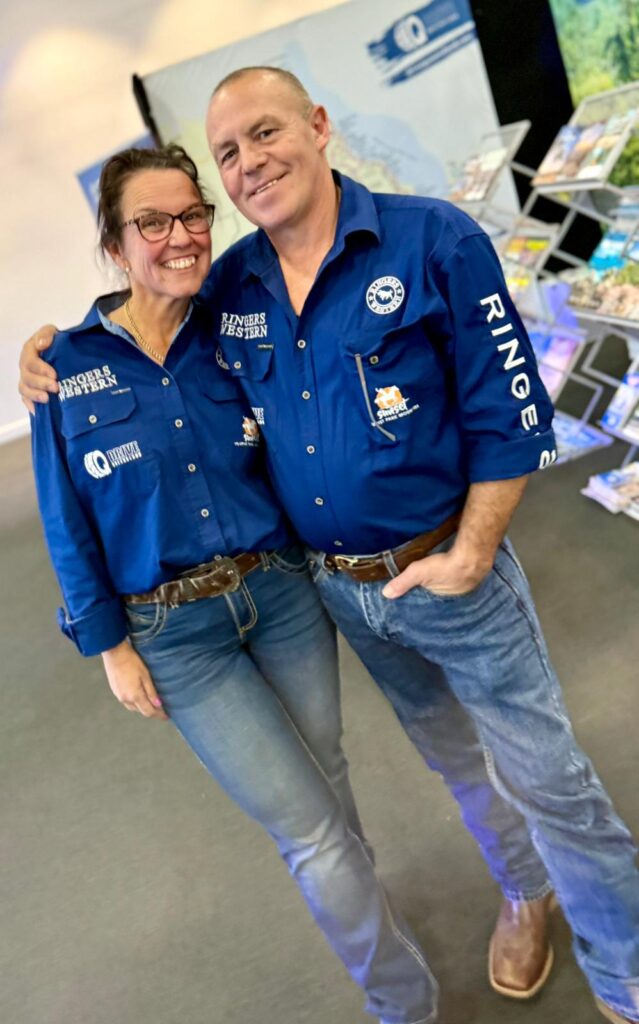 Two people wearing matching blue shirts and jeans stand together, smiling, with exhibition stands and brochures visible in the background.