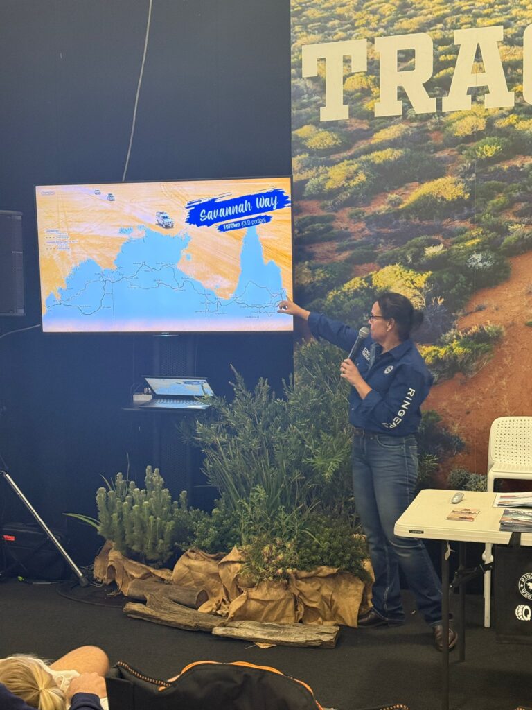 A woman in a blue shirt points to a map of the Savannah Way on a screen during a presentation, with plants and travel materials displayed nearby.