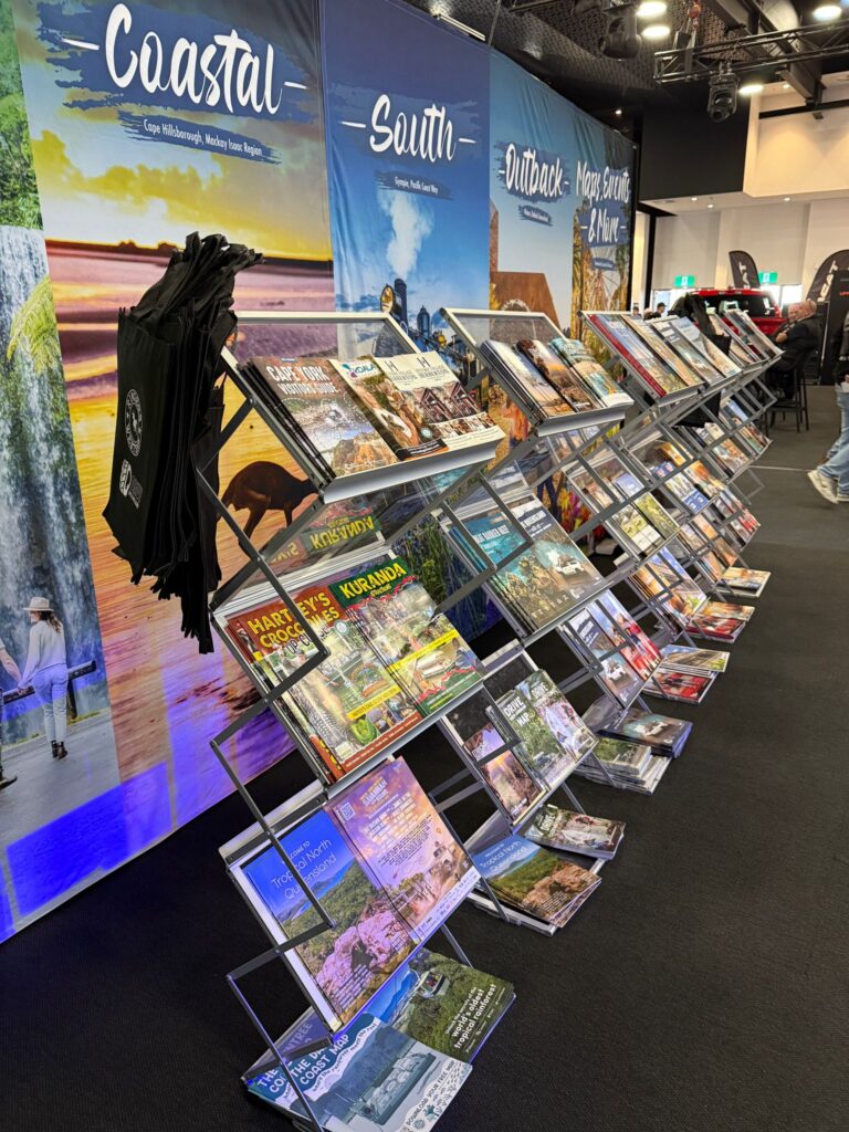 Multiple racks display travel brochures and magazines in front of large promotional banners labeled 