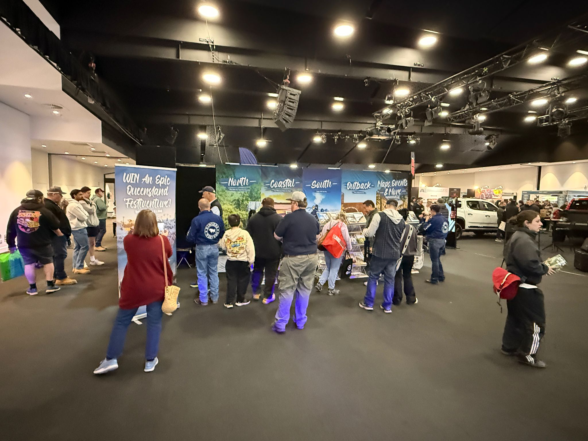 People stand in line indoors at an event, facing booths with large banners displaying travel destinations and information.