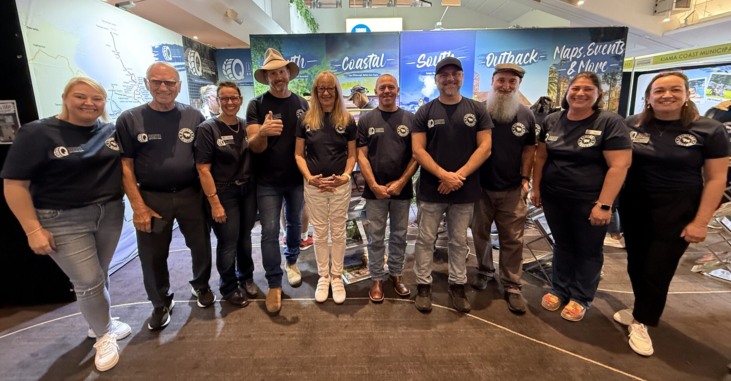 A group of eleven people wearing matching navy blue shirts stand together indoors at an event booth, smiling at the camera.
