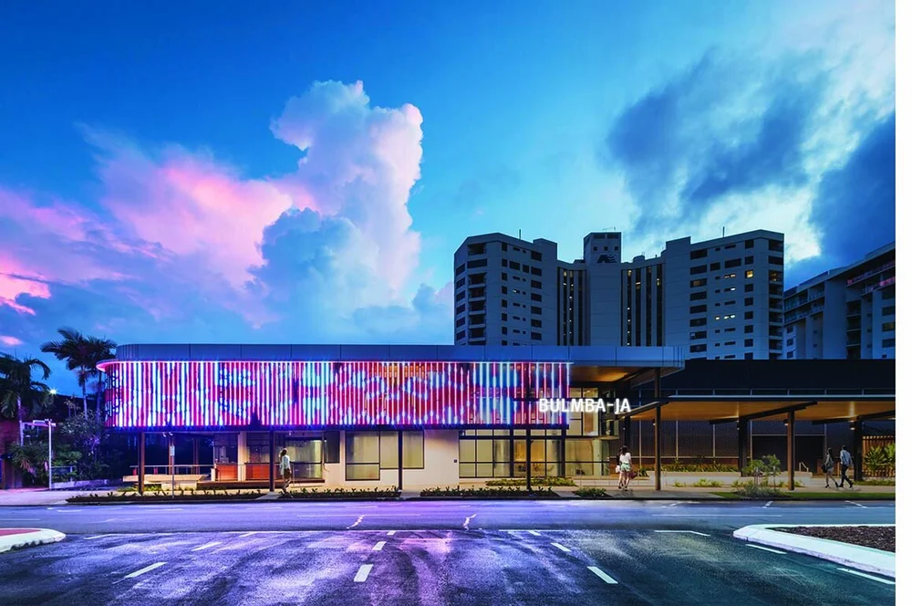 A modern building with colorful vertical light panels on the facade, "BULMBA-I A" signage, and high-rise apartments in the background at dusk.