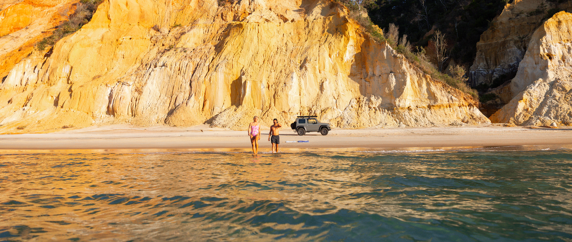 Two people stand on a sandy beach near a parked off-road vehicle, with large rocky cliffs in the background and water in the foreground.