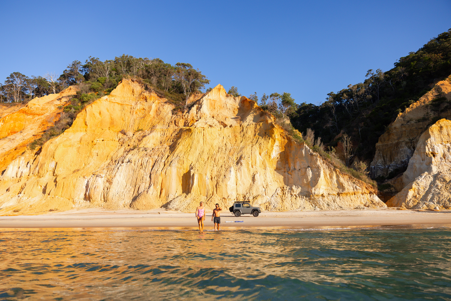 Two people stand on a sandy beach near a cliff with a parked vehicle under a clear blue sky, viewed from the water.