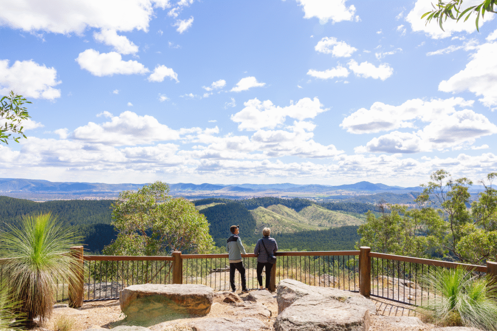 Two people stand at a fenced lookout point, overlooking a scenic landscape of hills, trees, and distant mountains under a blue sky with scattered clouds.