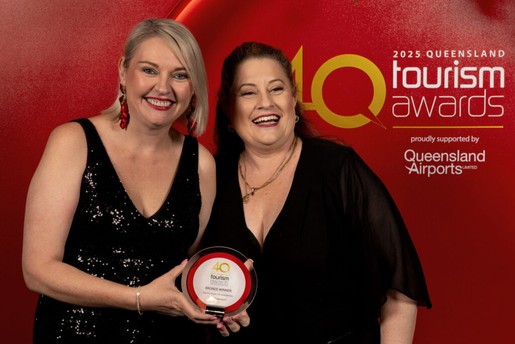 Two women in black dresses smile and hold a 2023 Queensland Tourism Awards plaque against a red backdrop featuring event branding and sponsor logos.