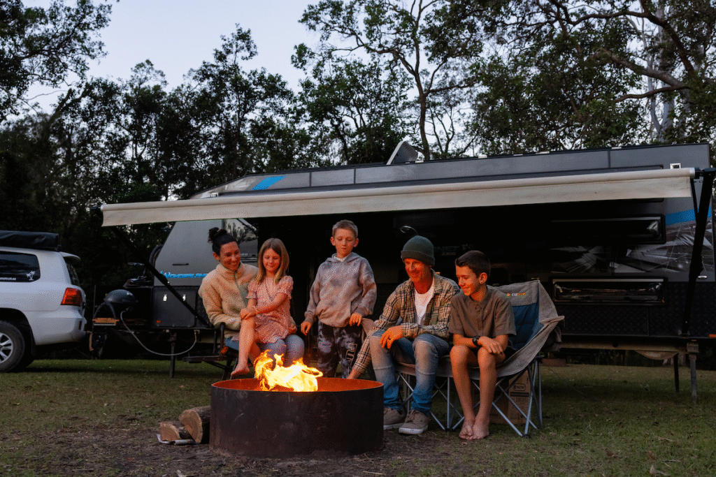 A family of five sits around a campfire in front of a camper trailer, with trees and a white vehicle in the background.