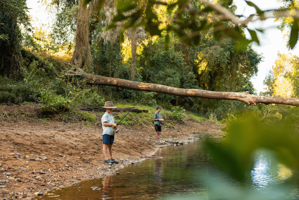 Two children stand on a riverbank holding fishing rods, surrounded by trees and greenery, with a fallen tree trunk in the background.