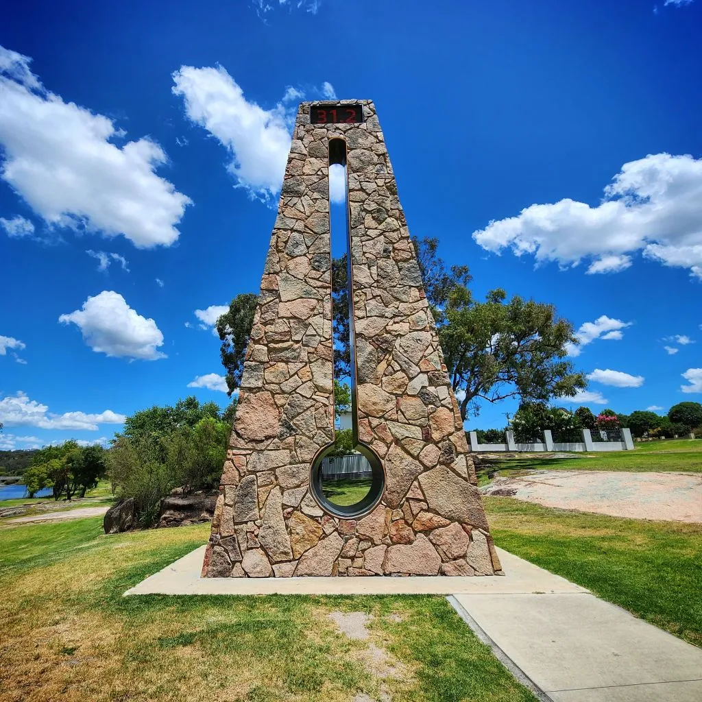 A tall, triangular stone monument with a vertical slit and circular hole at the base stands on a sidewalk in a grassy park under a blue sky with clouds.