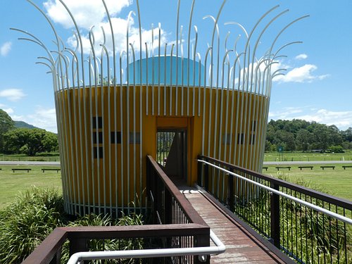 A yellow, circular building with vertical white metal rods on the roof, viewed from a ramped walkway, set in a grassy landscape under a blue sky.