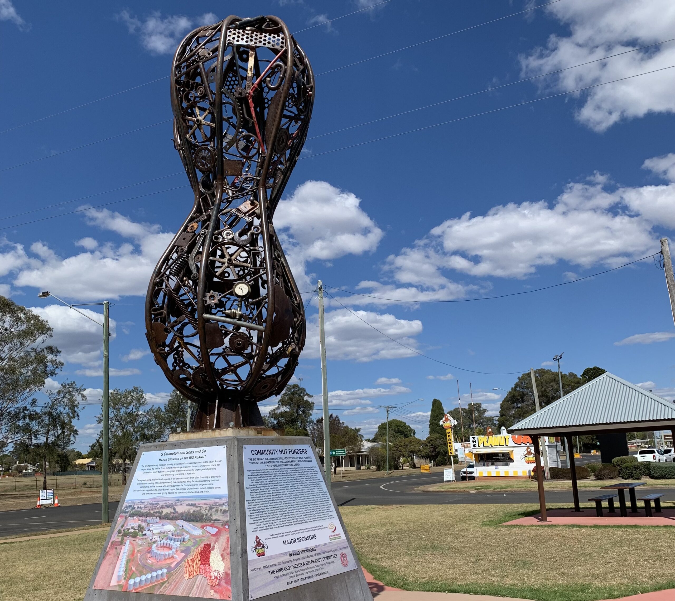 Large metal peanut-shaped sculpture stands on a concrete base with informational plaques in a grassy area; blue sky and scattered clouds in the background.
