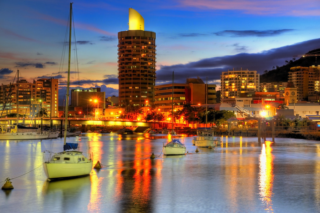 Several sailboats are docked in a calm harbor with city buildings and lights reflecting on the water at dusk.