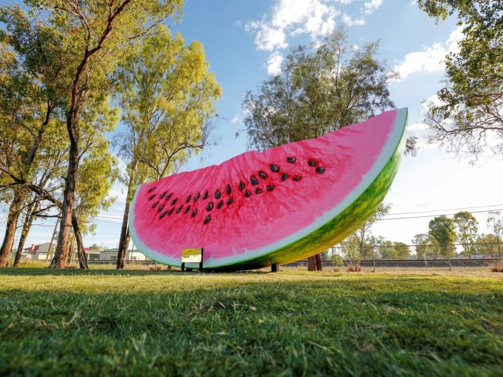 A large watermelon slice sculpture sits on a grassy field, surrounded by trees under a partly cloudy sky.
