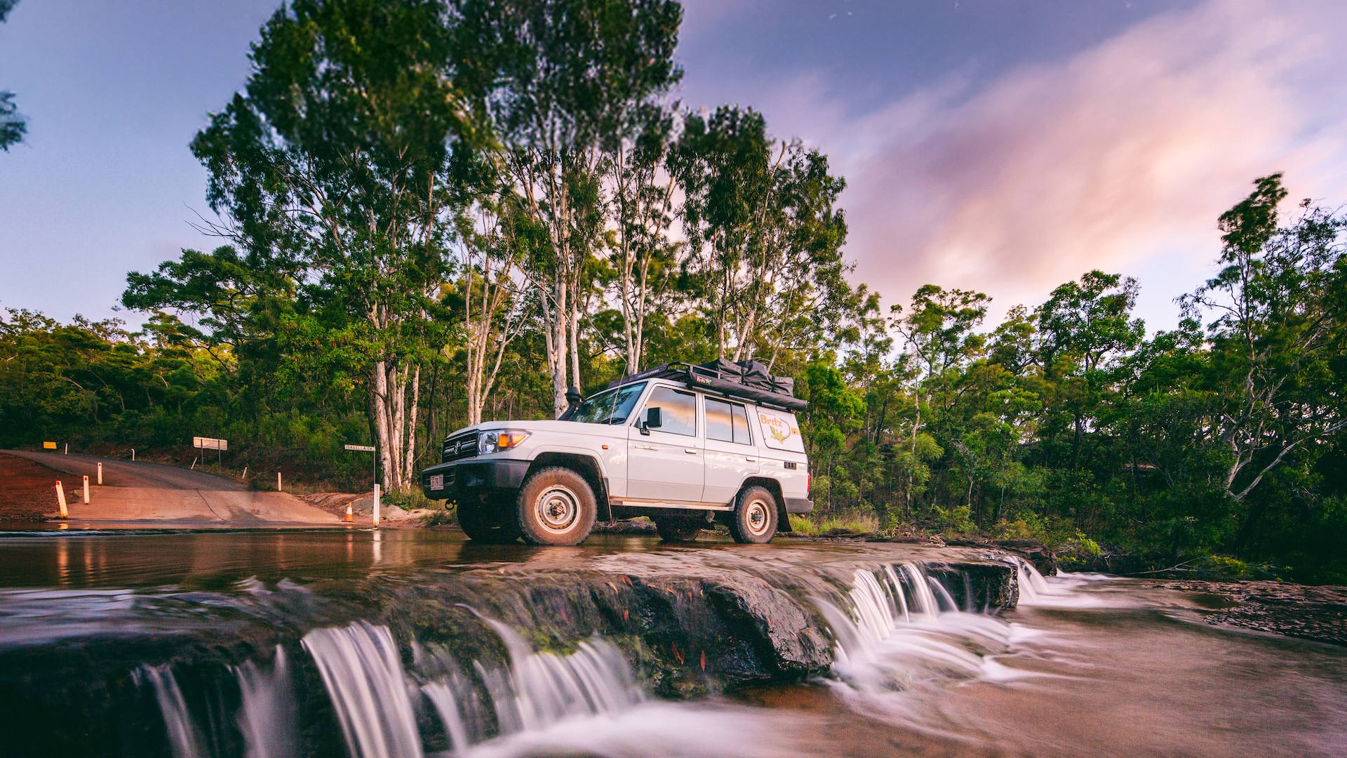 A white off-road vehicle is parked on a rocky ledge above a shallow waterfall, surrounded by trees under a partly cloudy sky at sunset.