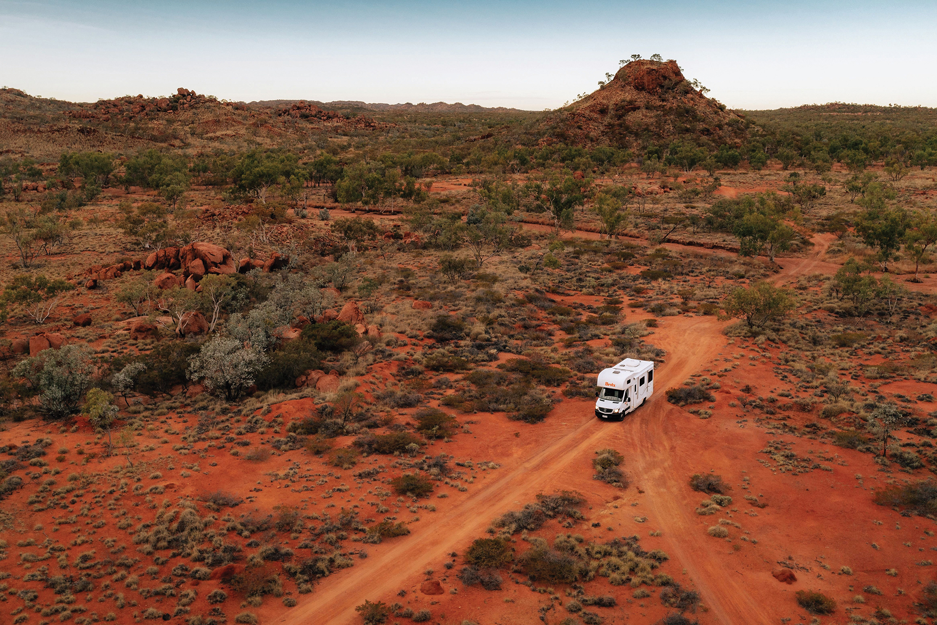 A white RV drives on a dirt road through a red, arid landscape with sparse vegetation and hills in the background.