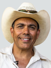 A middle-aged man wearing a white cowboy hat, sunglasses on top, and a light-colored shirt smiles at the camera against a plain background.