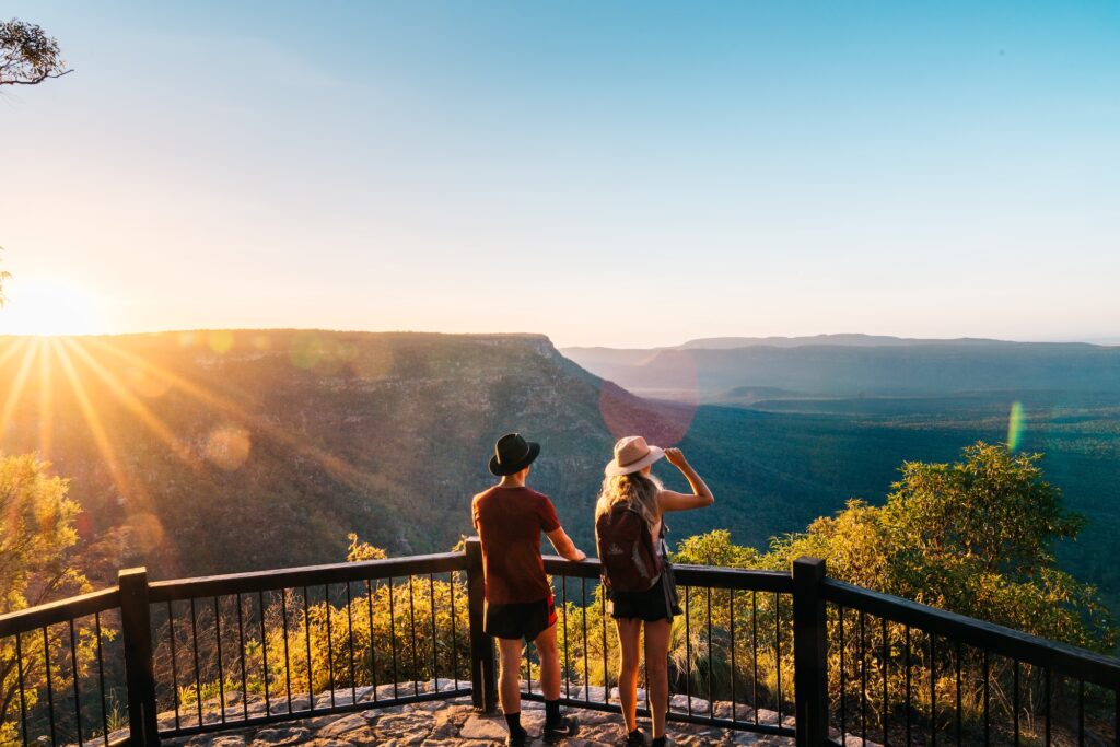 Two people stand on a fenced lookout overlooking a scenic valley and plateaus at sunset, with sunlight streaming over the landscape.