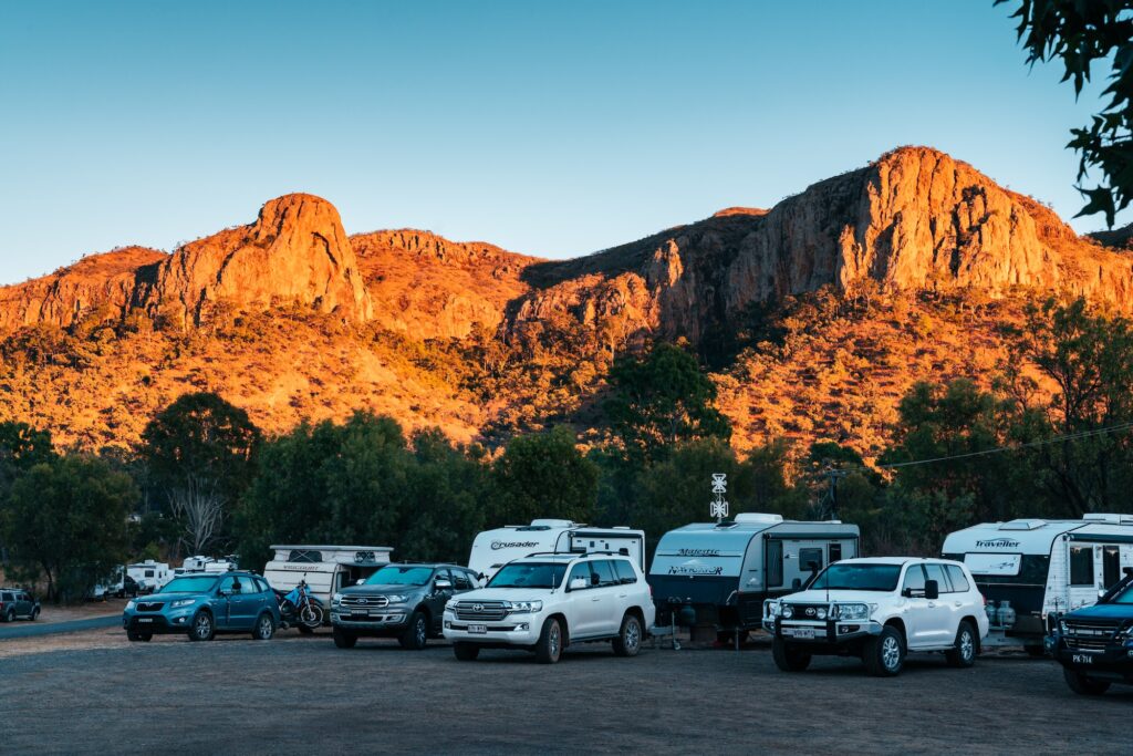 Several cars and campervans are parked in a lot at the base of rocky, sunlit hills with trees scattered around.