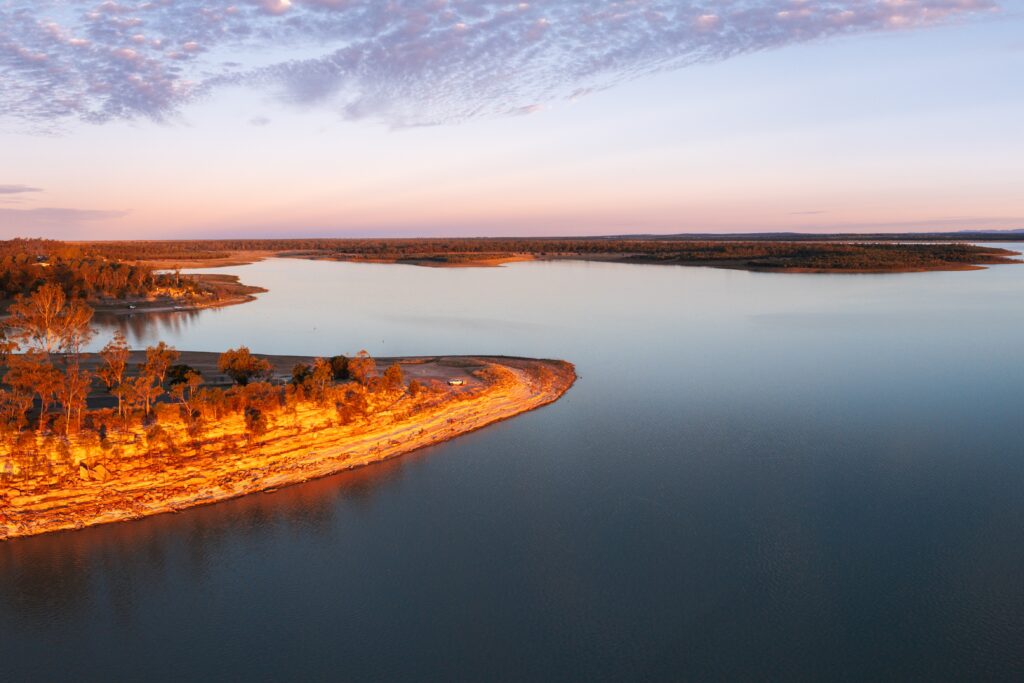 A wide river or lake with calm water curves around a peninsula with trees under a partly cloudy sky at sunset.
