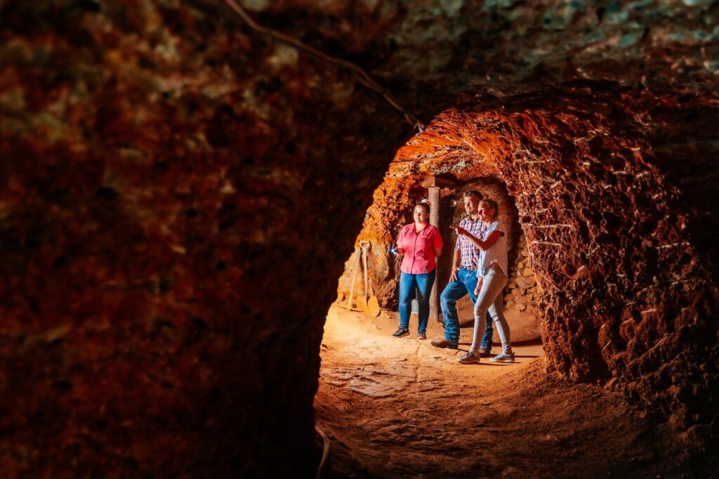 Three people stand inside a reddish-brown, dimly lit underground tunnel with uneven walls and ceiling.