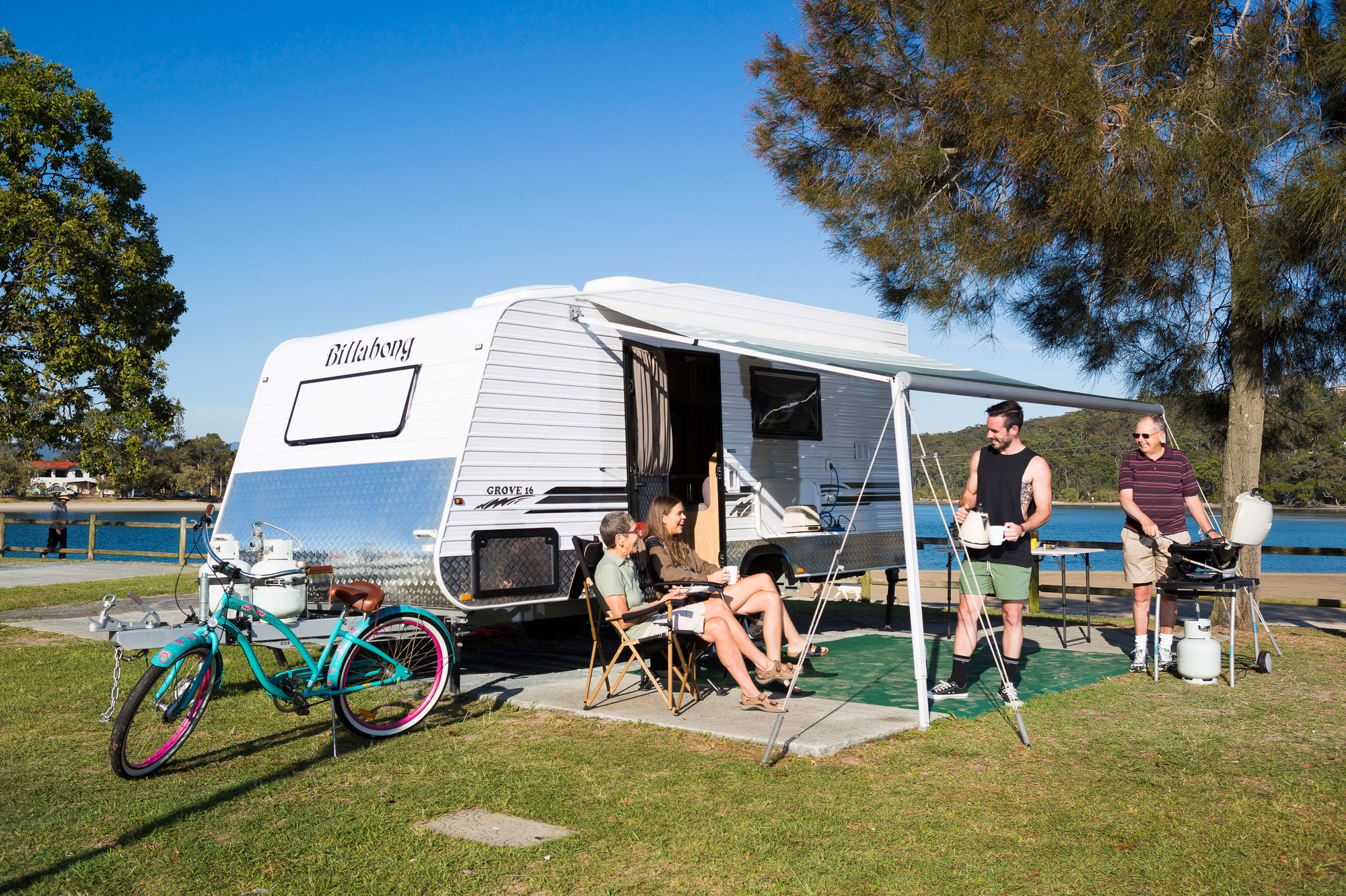 Four people gather outside a white camper van by a lake, with a barbecue grill, bicycles, chairs, and a tree nearby on a sunny day.