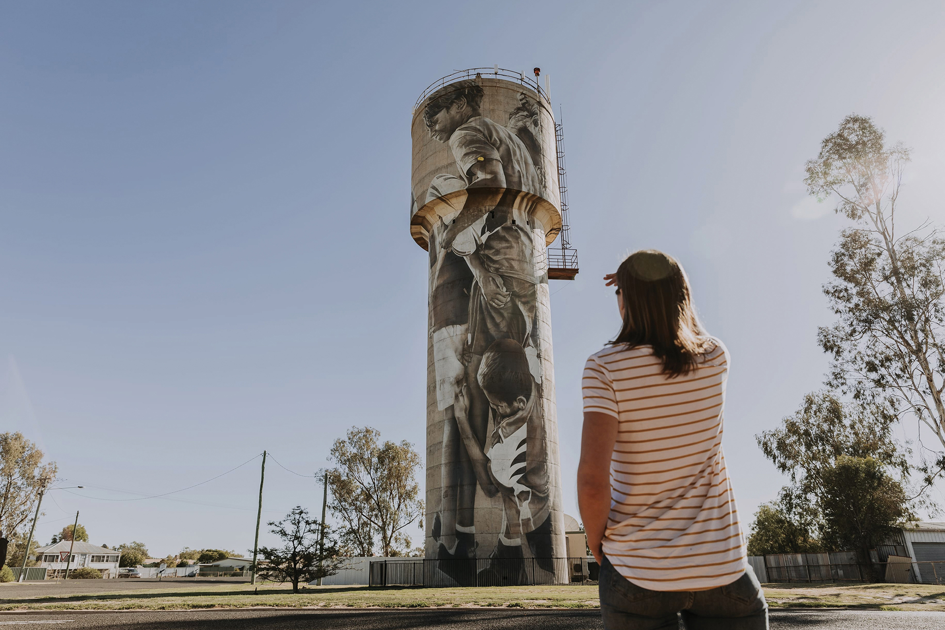 A person wearing a striped shirt and cap stands in front of a tall water tower painted with a large mural of people and hands, on a clear day.