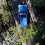 A blue car drives along a dirt road surrounded by trees, viewed from above.