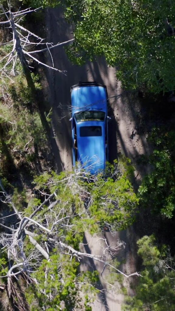 A blue car drives along a dirt road surrounded by trees, viewed from above.