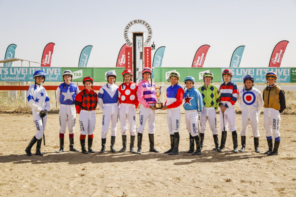 Twelve jockeys in colorful uniforms stand in a row holding a trophy at a racetrack with banners and flags in the background.
