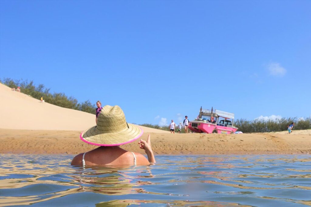 Person in a straw hat wades in shallow water near a sandy shore with a pink boat and several people in the background under a clear blue sky.