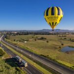 A yellow and black hot air balloon floats over a rural landscape with fields, a pond, and a highway on a clear day.