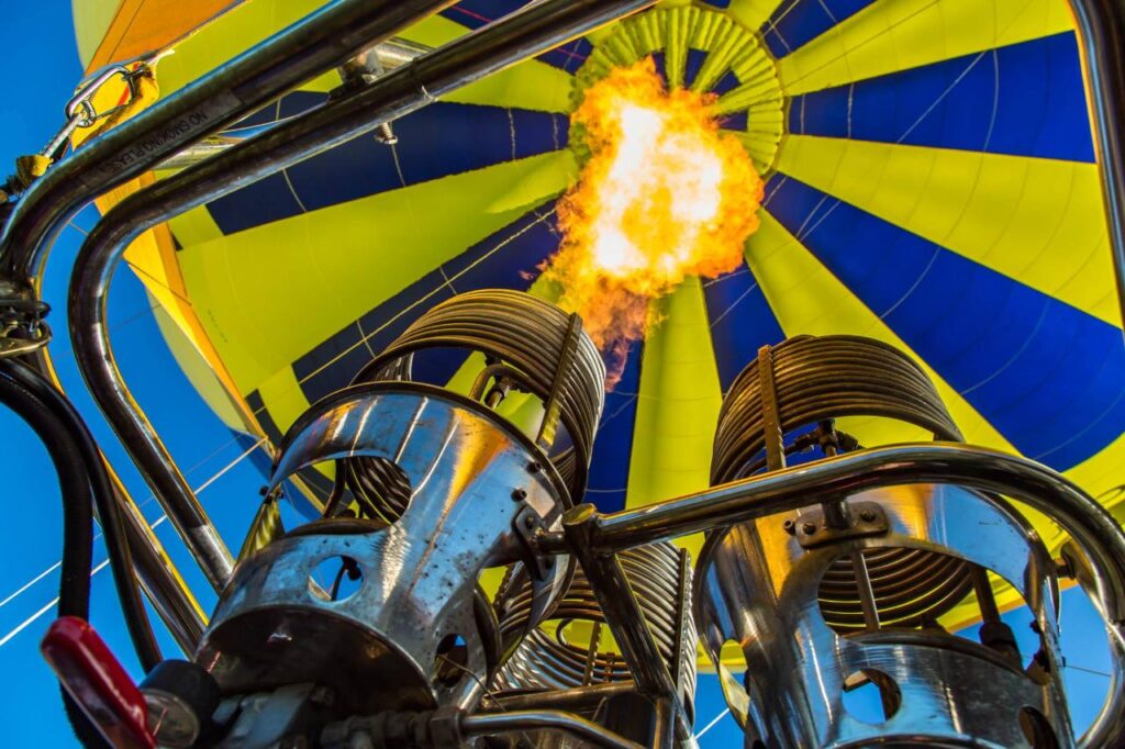 Close-up view from below of a hot air balloon burner shooting a flame upward into a yellow and blue balloon envelope.