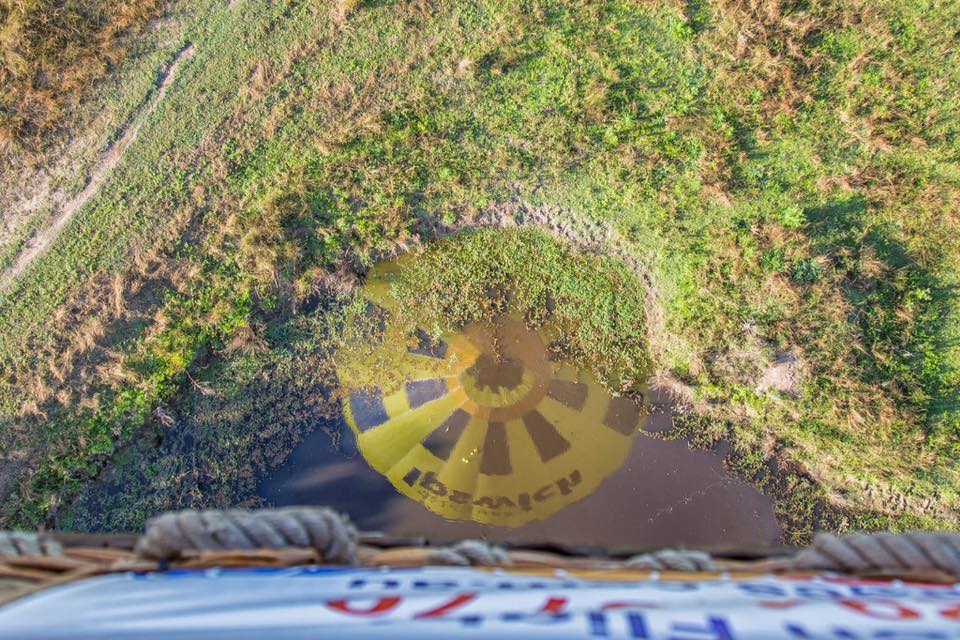 A hot air balloon casts a round yellow shadow with dark sections on a grassy and marshy area, viewed from the basket above.