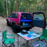 A pink SUV with its trunk open is parked at a forest campsite, with camping gear, a table, camping stove, chairs, and supplies set up nearby among tall trees.