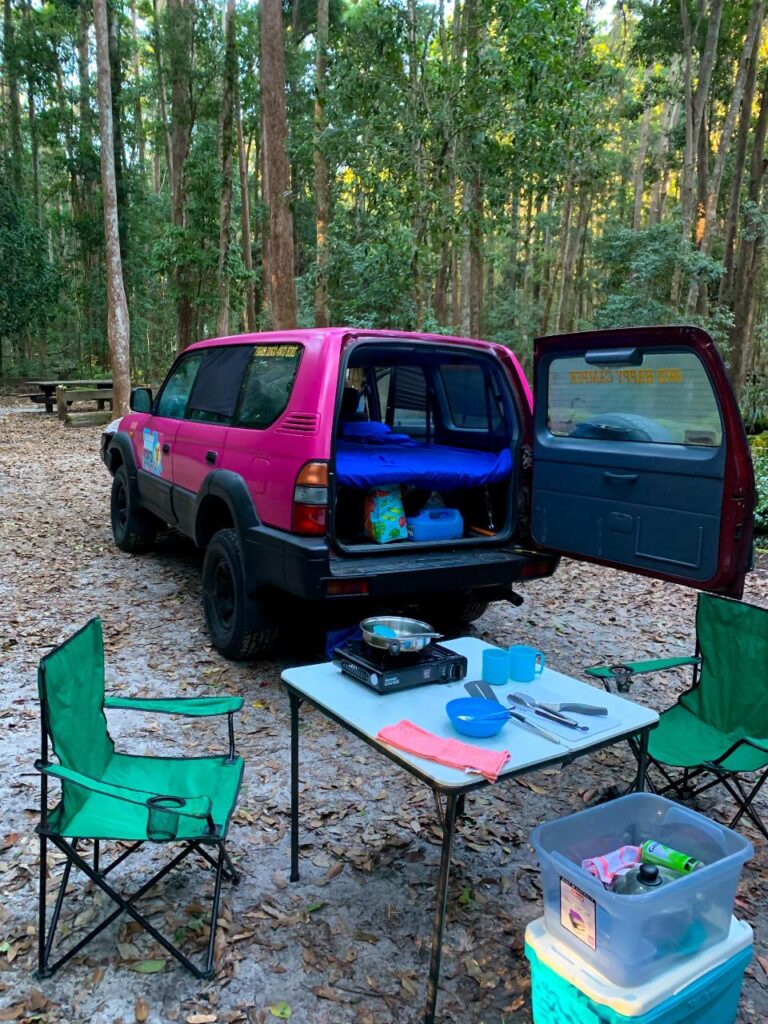 A pink SUV with its trunk open is parked at a forest campsite, with camping gear, a table, camping stove, chairs, and supplies set up nearby among tall trees.