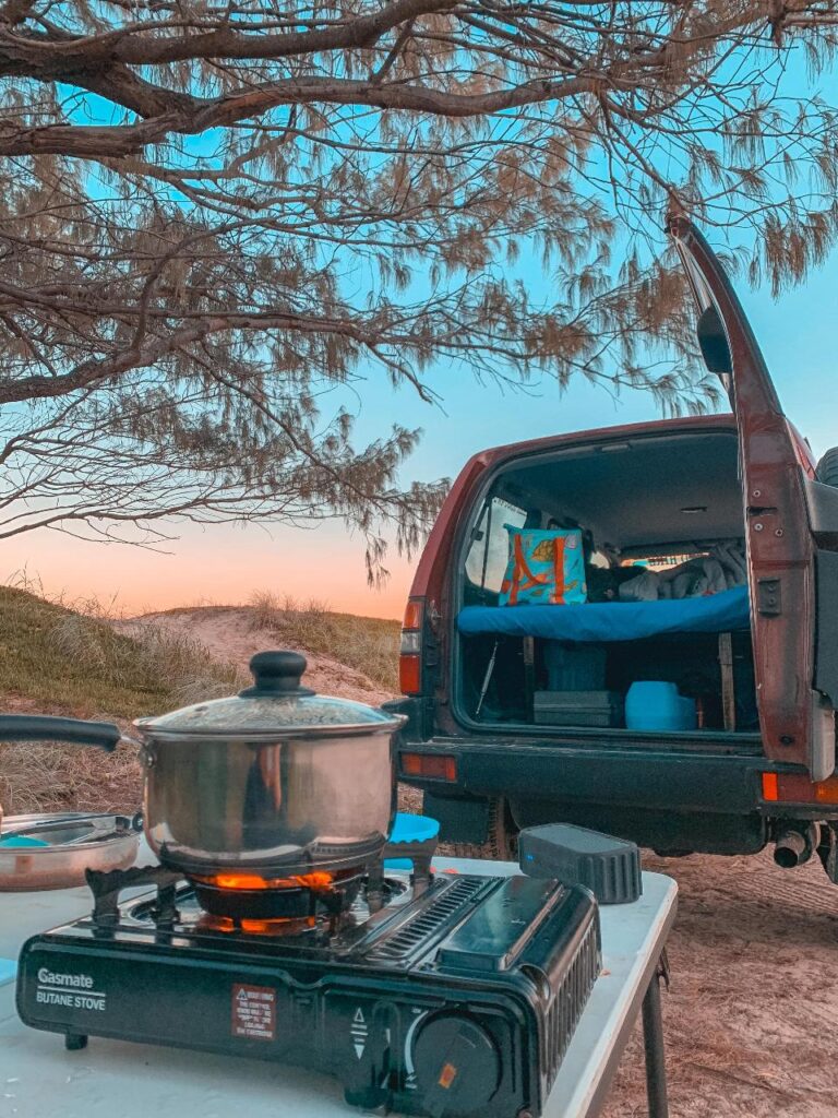 A butane stove with a pot sits on a table near a parked vehicle with its back door open, showing a bed setup inside. Trees and sky are visible in the background.