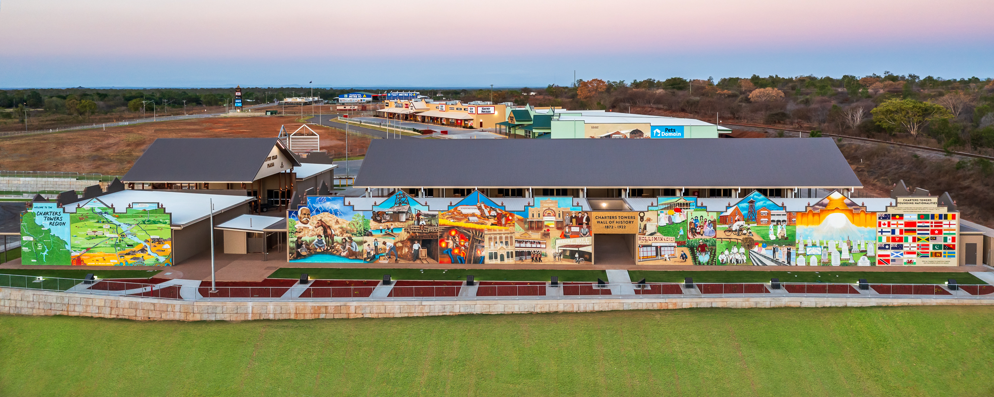 A school building with colorful murals on its exterior walls, set against a grassy field and a backdrop of trees and additional buildings at sunset.