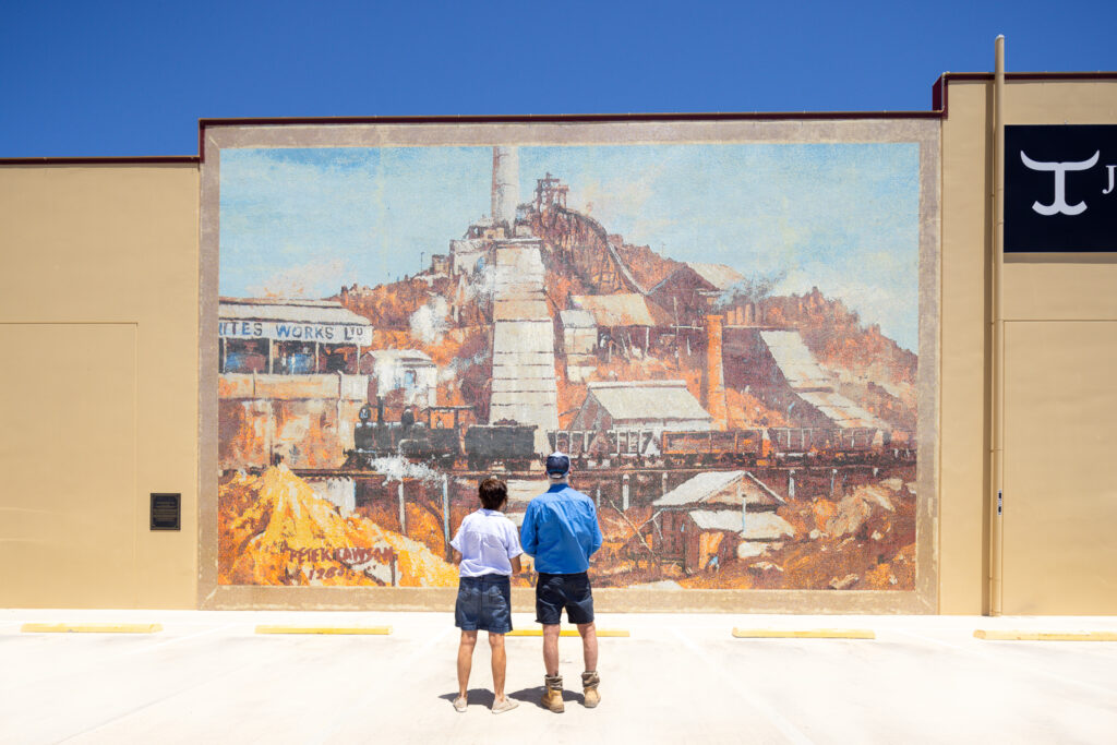 Two people stand in an empty parking lot, facing a mural on a beige wall depicting an industrial mining scene with buildings and a train.