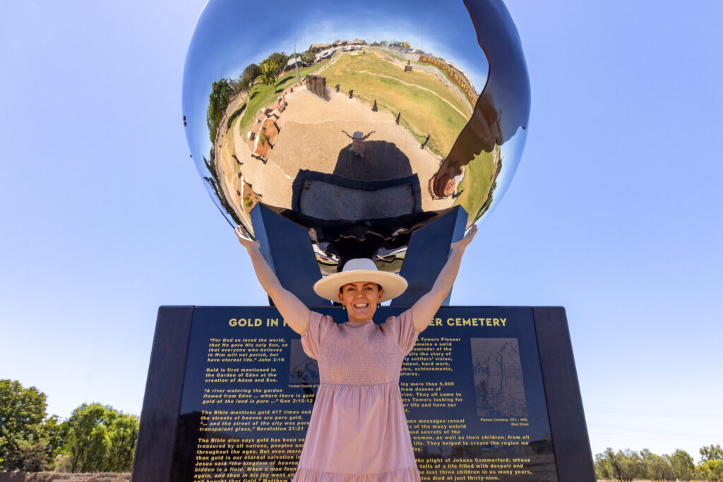 A woman in a pink dress and white hat poses with arms raised in front of a large reflective sphere at a historical cemetery site on a sunny day.
