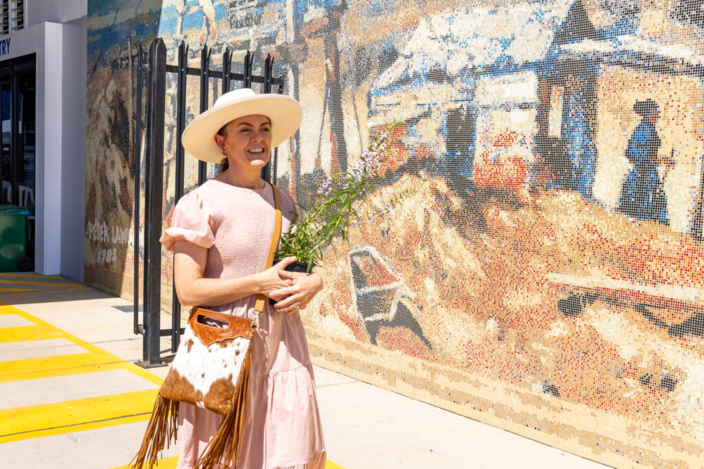 A woman in a pink dress and wide-brimmed hat stands holding flowers in front of a mural, with a brown leather bag slung over her shoulder.