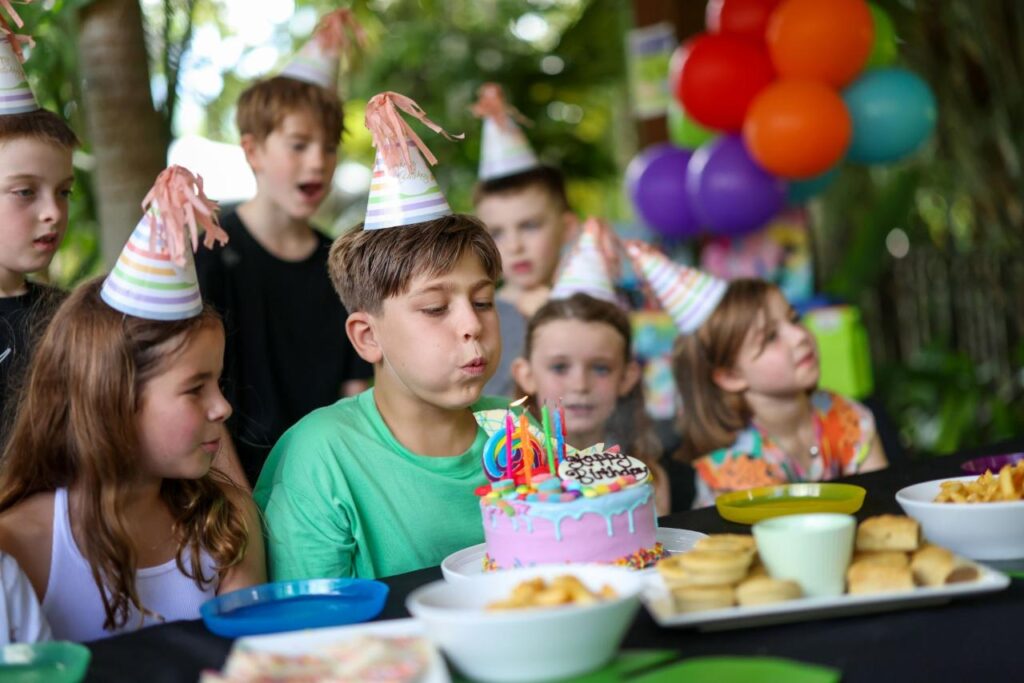 A boy in a green shirt blows out candles on a birthday cake, surrounded by children in party hats at a table with food and colorful balloons in the background.