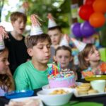 A boy in a green shirt blows out candles on a birthday cake, surrounded by children in party hats at a table with food and colorful balloons in the background.