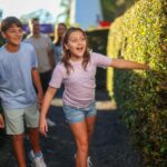 Two children walk through a hedge maze, smiling and looking ahead, while two adults follow behind them on a sunny day.
