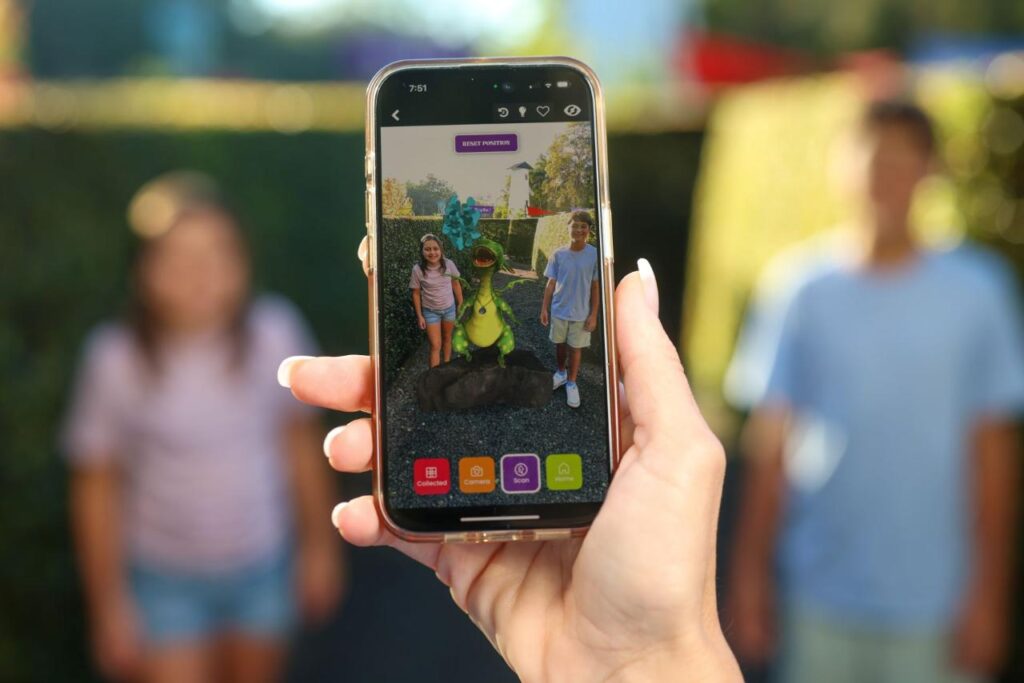 A person holds a smartphone taking a photo of two people standing next to a cartoon dinosaur digitally added via augmented reality.