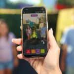 A person holds a smartphone taking a photo of two people standing next to a cartoon dinosaur digitally added via augmented reality.