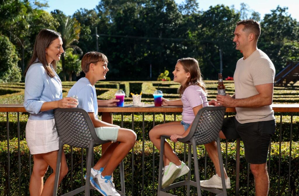Four people, two adults and two children, sit and stand around an outdoor bar table with drinks, surrounded by greenery and sunlight.