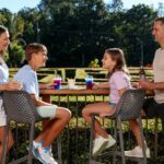 Four people, two adults and two children, sit and stand around an outdoor bar table with drinks, surrounded by greenery and sunlight.