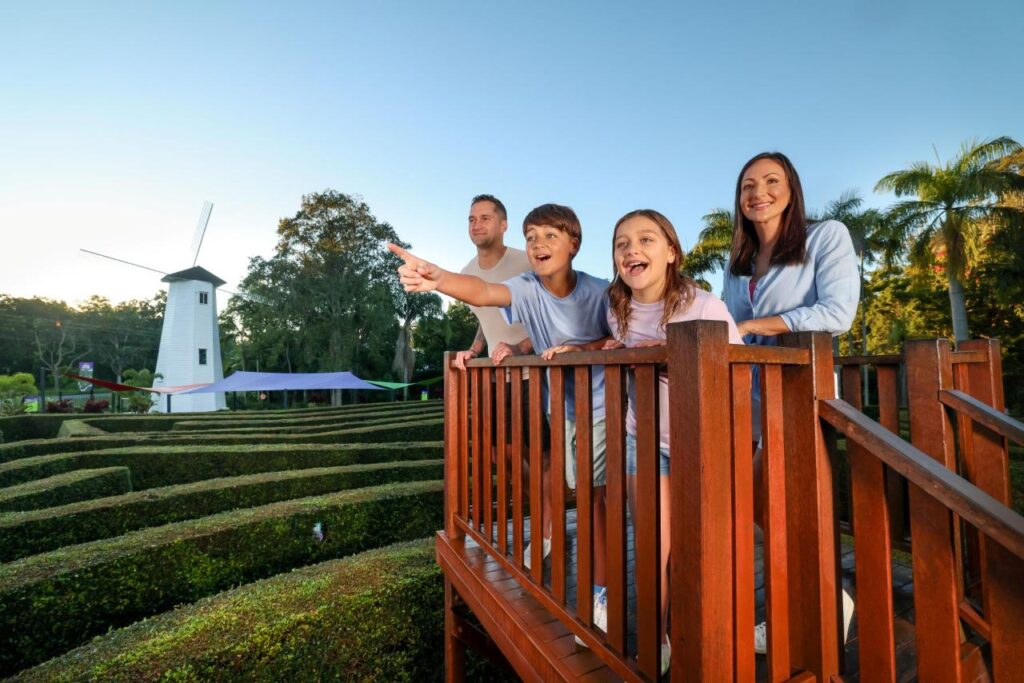 A family of four stands on a wooden platform overlooking a hedge maze, with a white windmill and trees in the background under a clear sky.